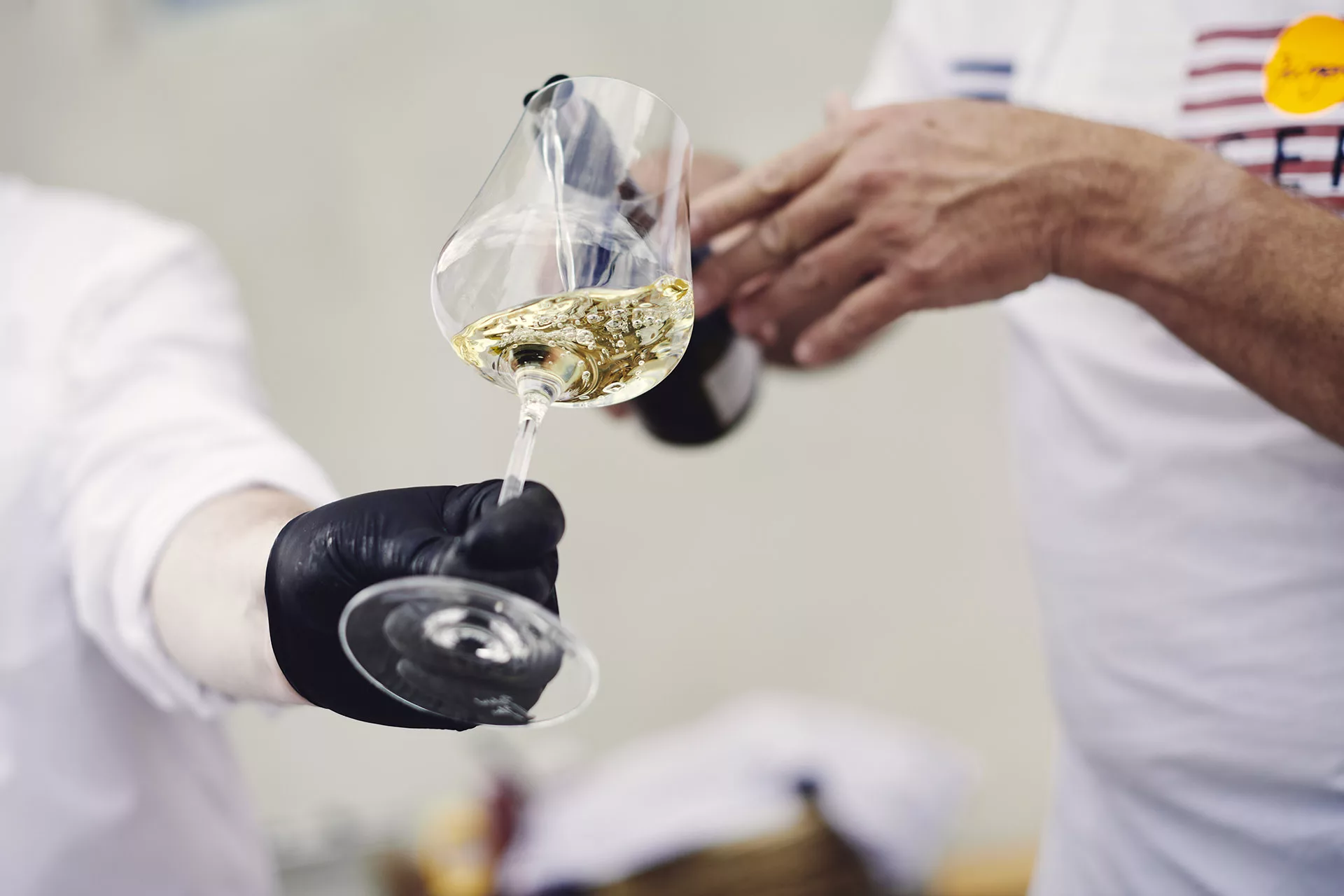 Person mit schwarzem Handschuh hält ein Glas, in das Weißwein eingeschenkt wird.