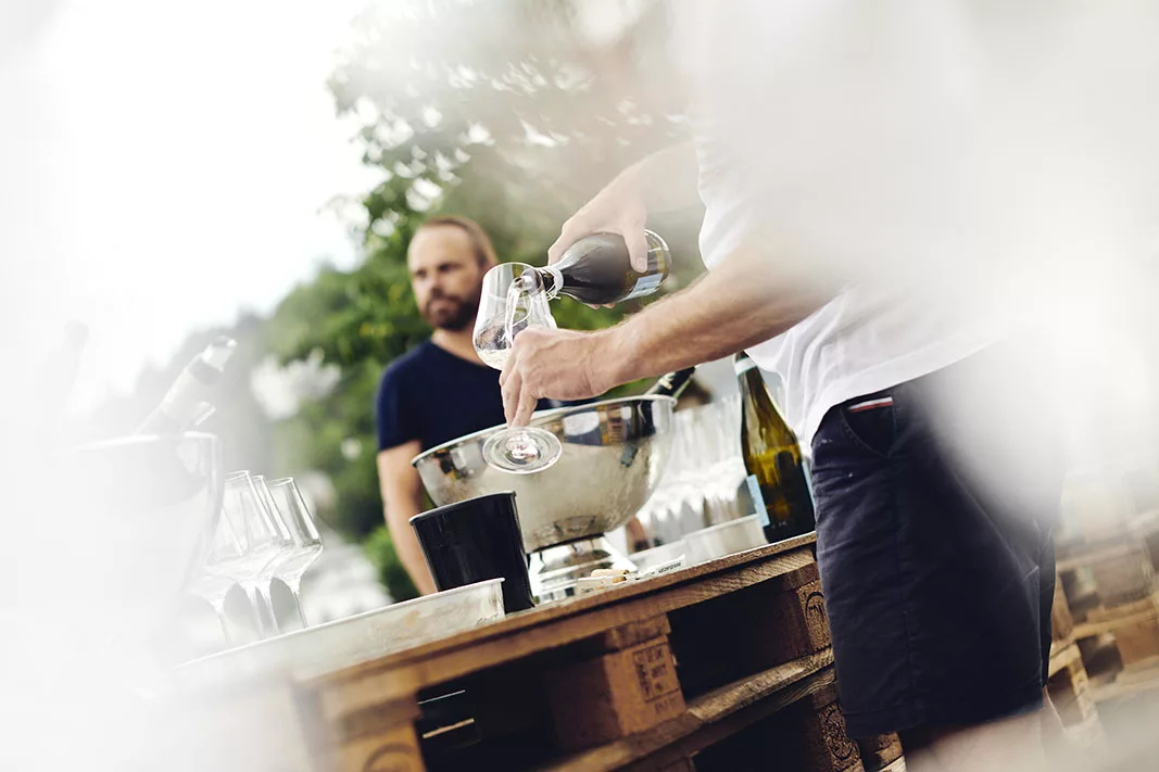 Person schenkt Wein in Glas bei einer Outdoor-Veranstaltung ein; Flaschen und Gläser auf Holztischen.