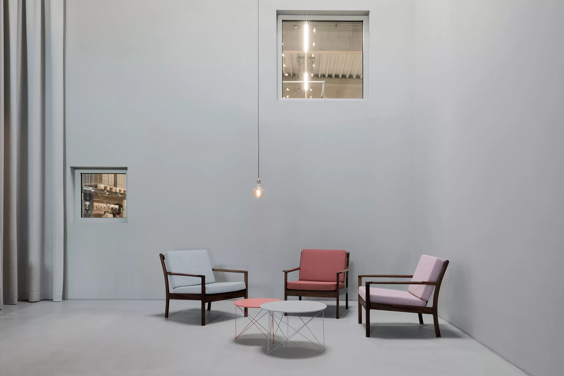Drei Stühle in Pastellfarben um kleine Tische und eine hängende Glühbirne vor grauer Wand, Fenster im Hintergrund.