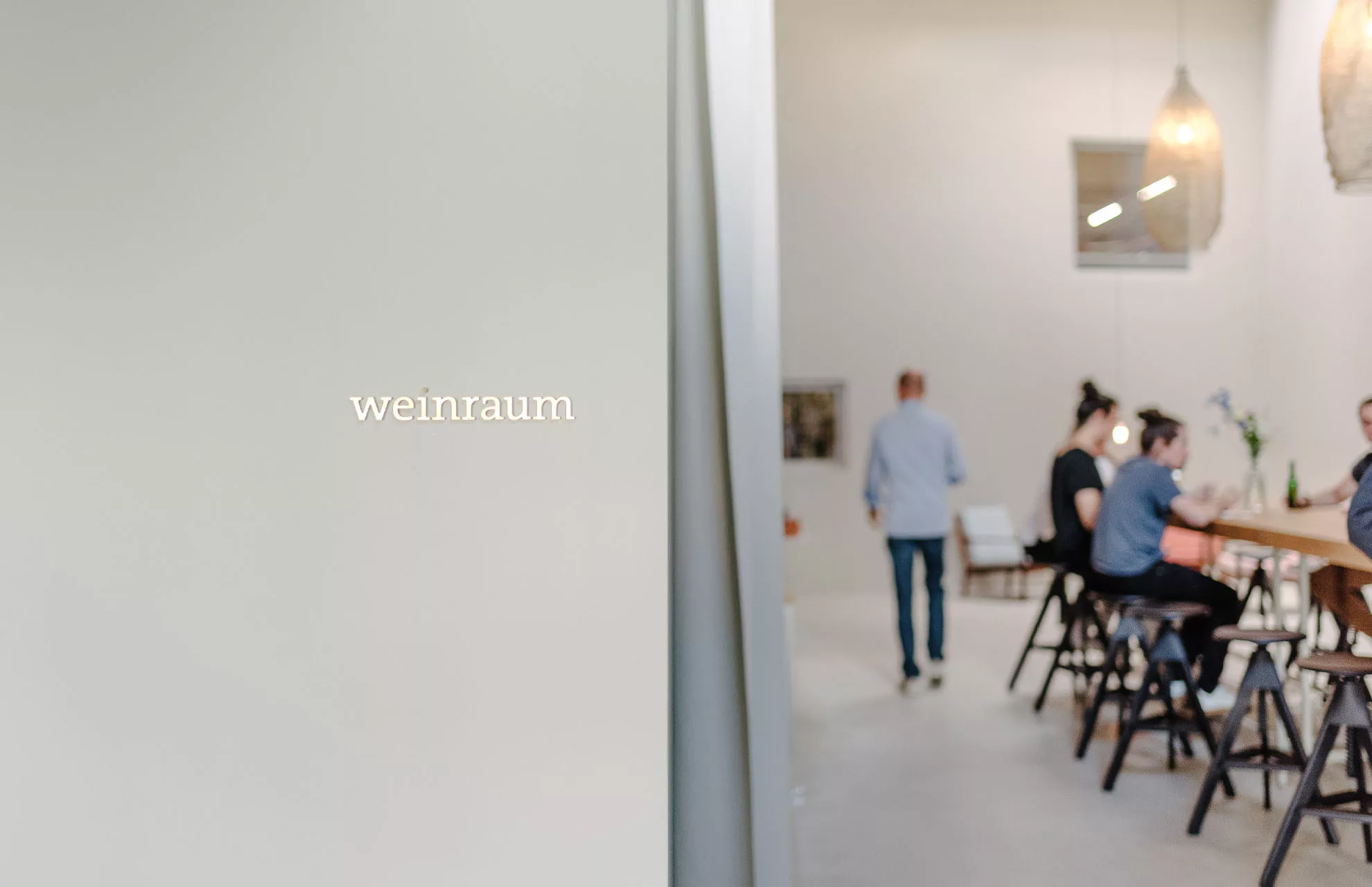 Raum mit dem Schriftzug weinraum, Personen sitzen an einem Tisch, entspannte Atmosphäre, helle Beleuchtung.