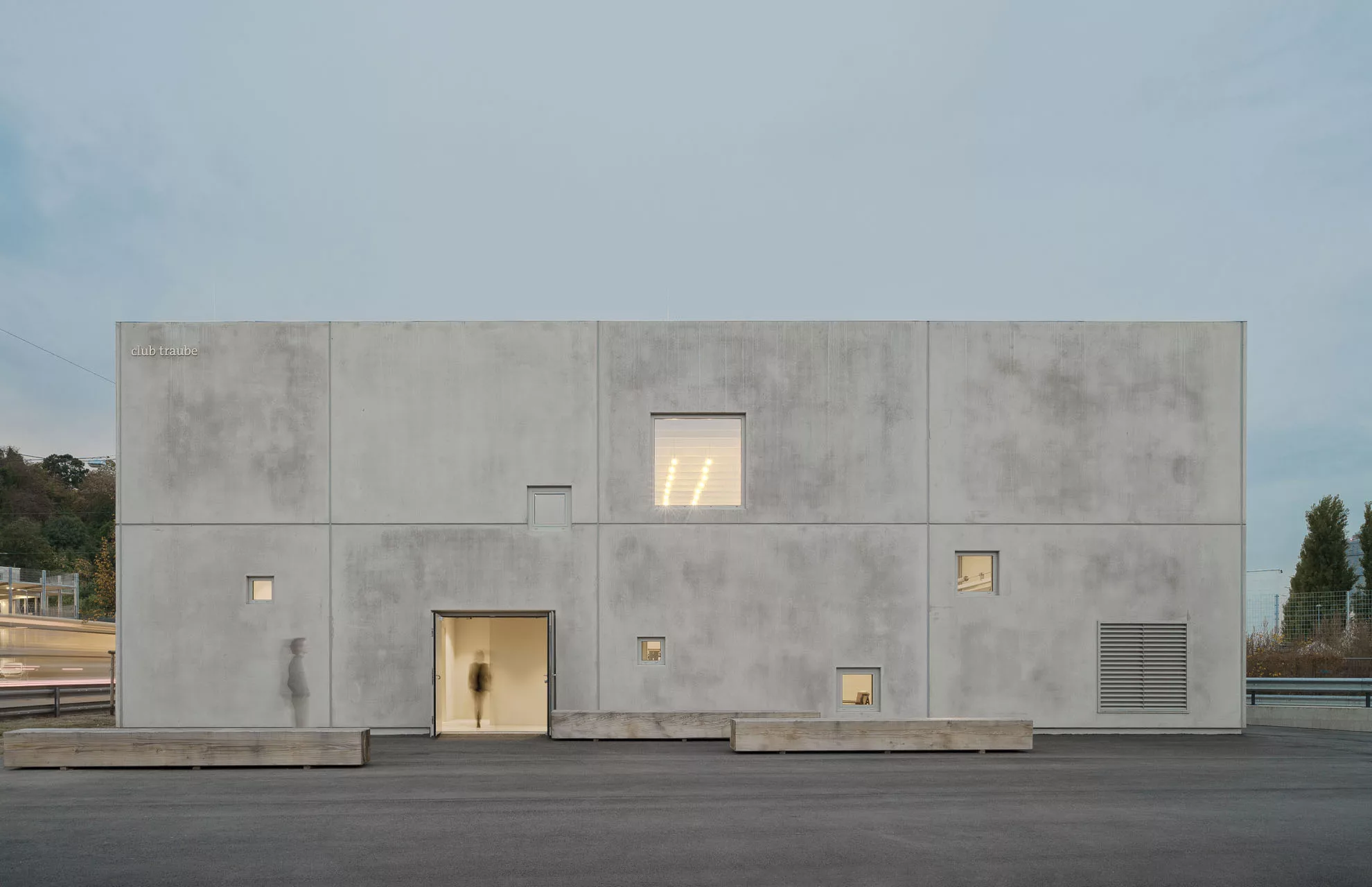 Modernes, graues Gebäude mit mehreren Fenstern und Tür, person vor Eingang verschwommen, zu sehen im Hintergrund Bäume.