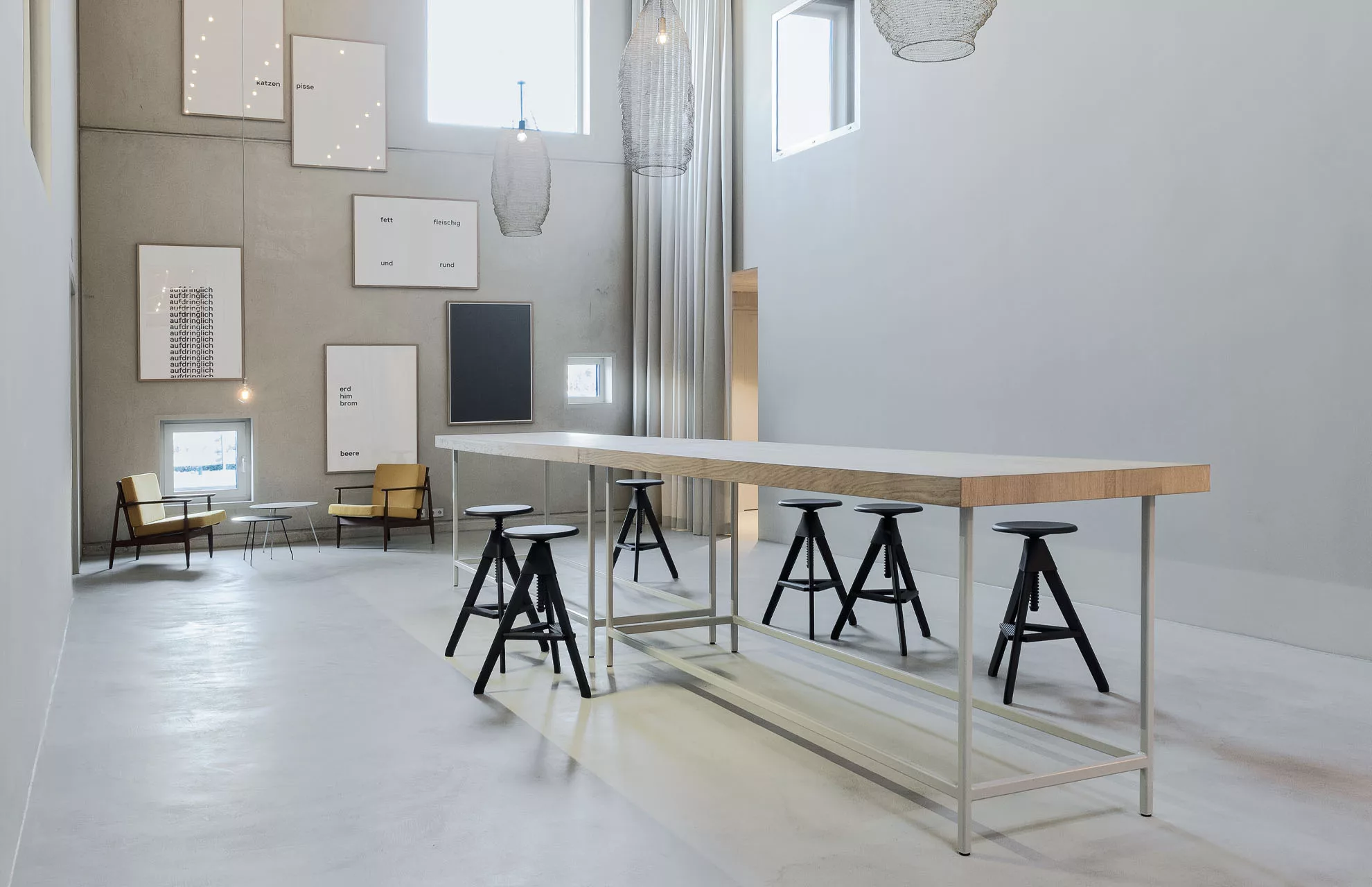 Moderner Raum mit hohem Tisch, schwarzen Hockern und Kunstwerken an der Betonwand. Zwei Sessel neben kleinen Tischen.
