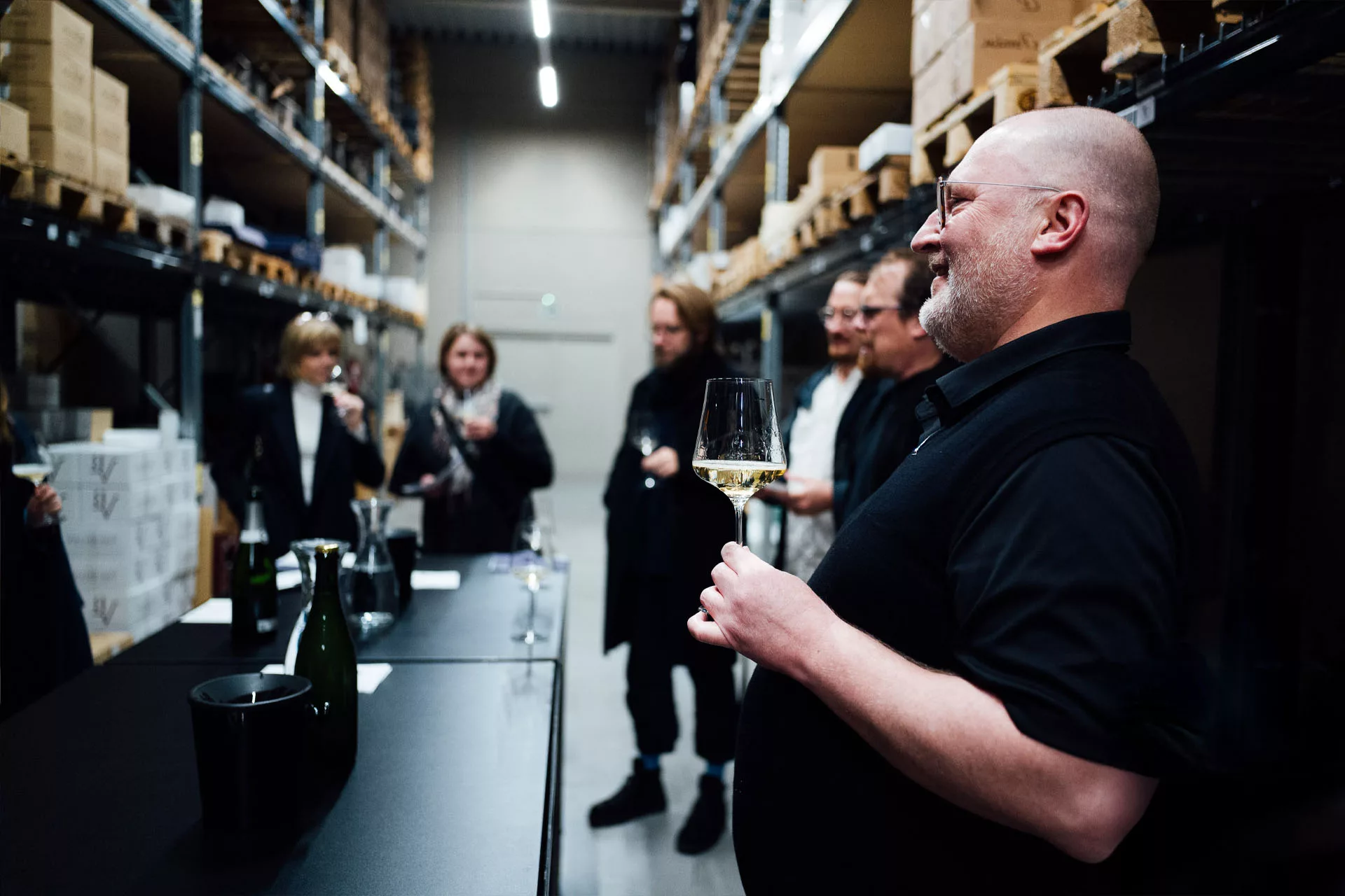 Mehrere Personen verkosten Wein in einem Lager mit Regalen voller Kartons und betrachten einander.