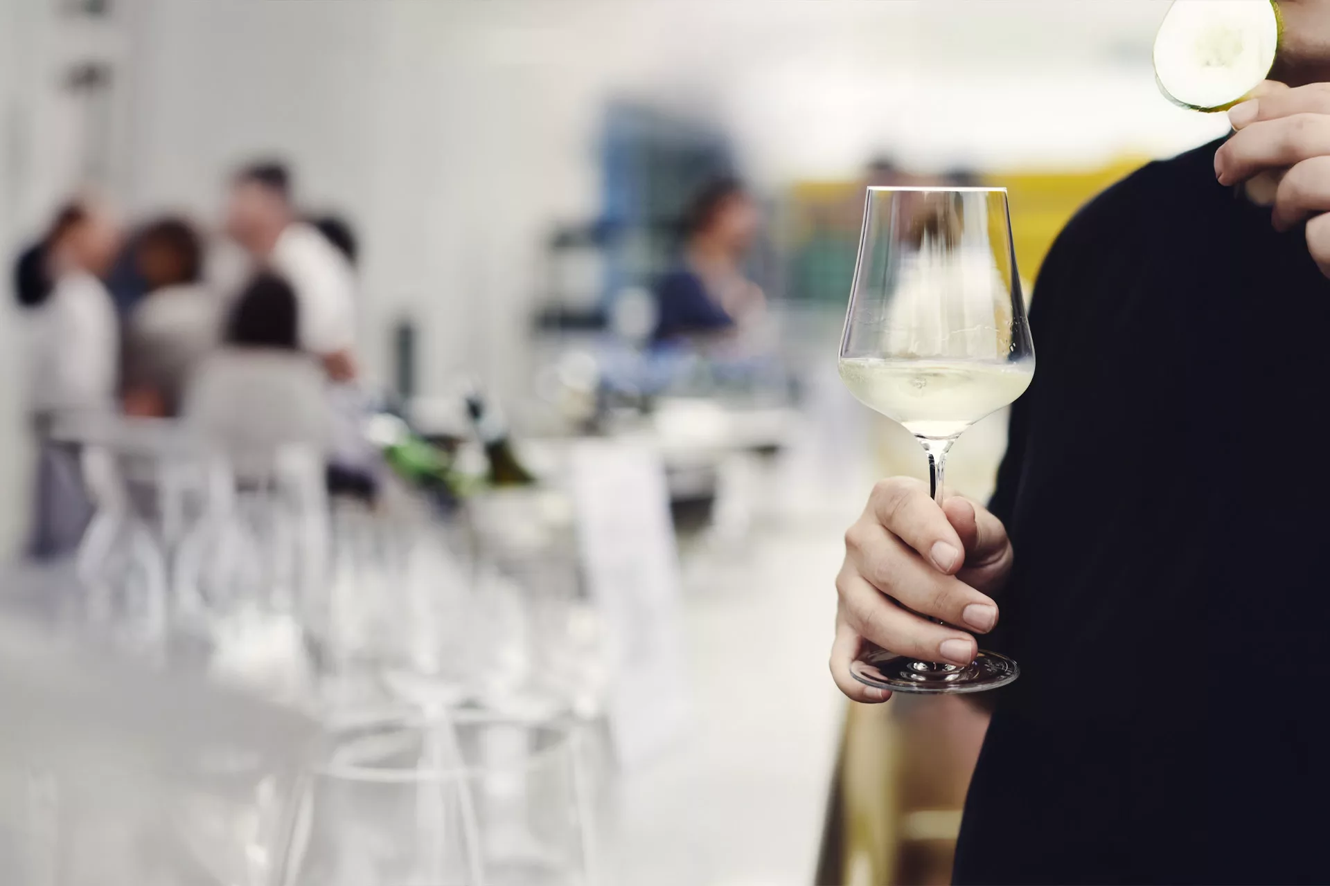 Person hält ein Glas Weißwein und eine Gurkenscheibe in einer Bar, im Hintergrund verschwommene Menschen.