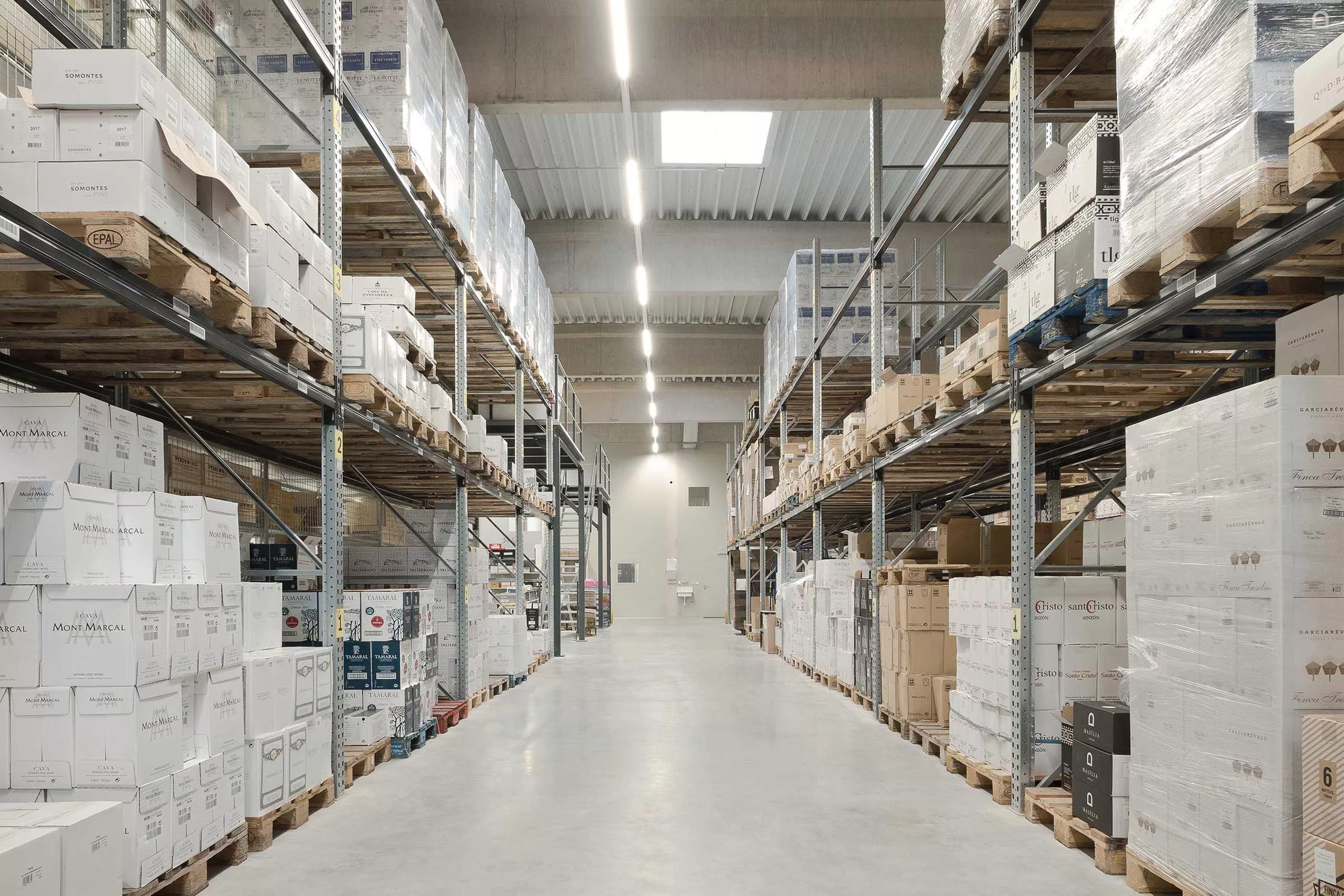 Regalreihen in einem Lager mit gestapelten Kartons auf Paletten unter hellem Deckenlicht.