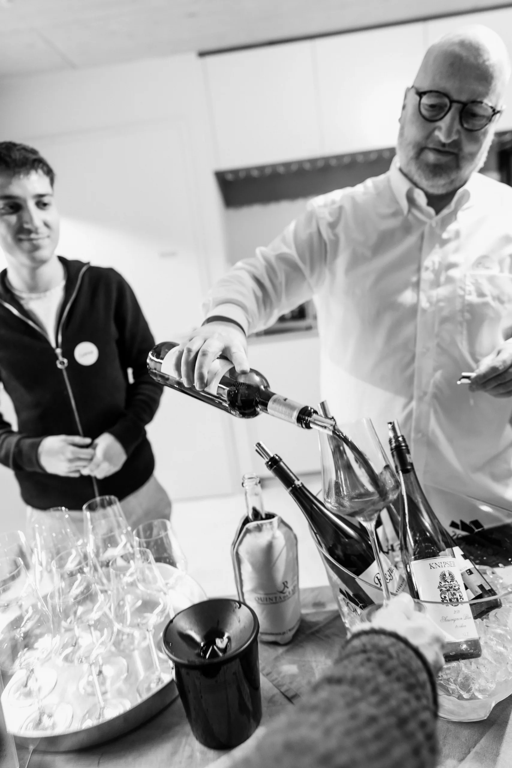 Mann schenkt Wein in Glas ein, während ein anderer zusieht. Tisch mit Flaschen und Gläsern in einem modernen Raum.