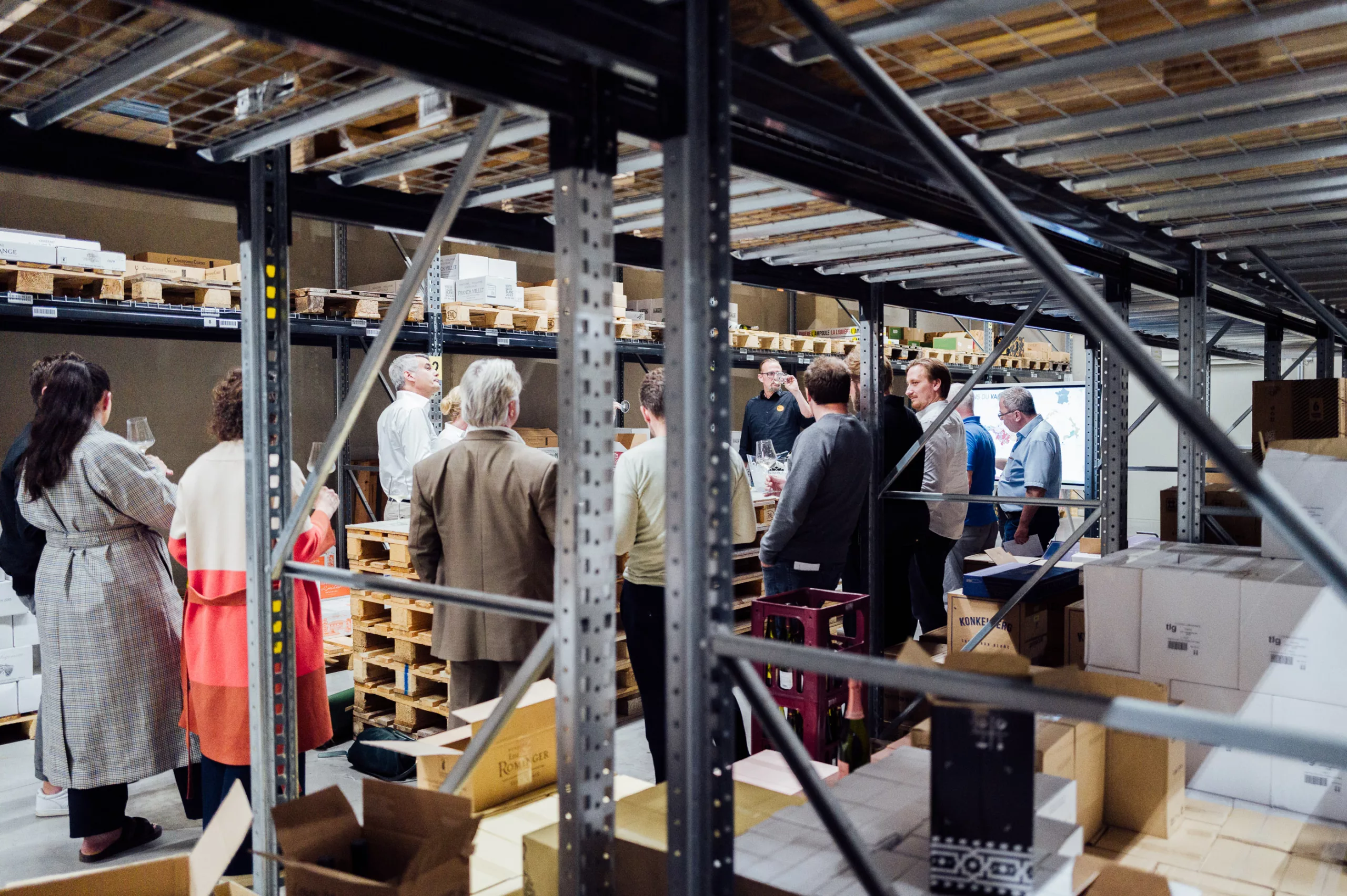 Eine Gruppe von Menschen steht in einem Lager zwischen Regalen und Kartons, einige halten Gläser, als ob sie an einer Führung teilnehmen.