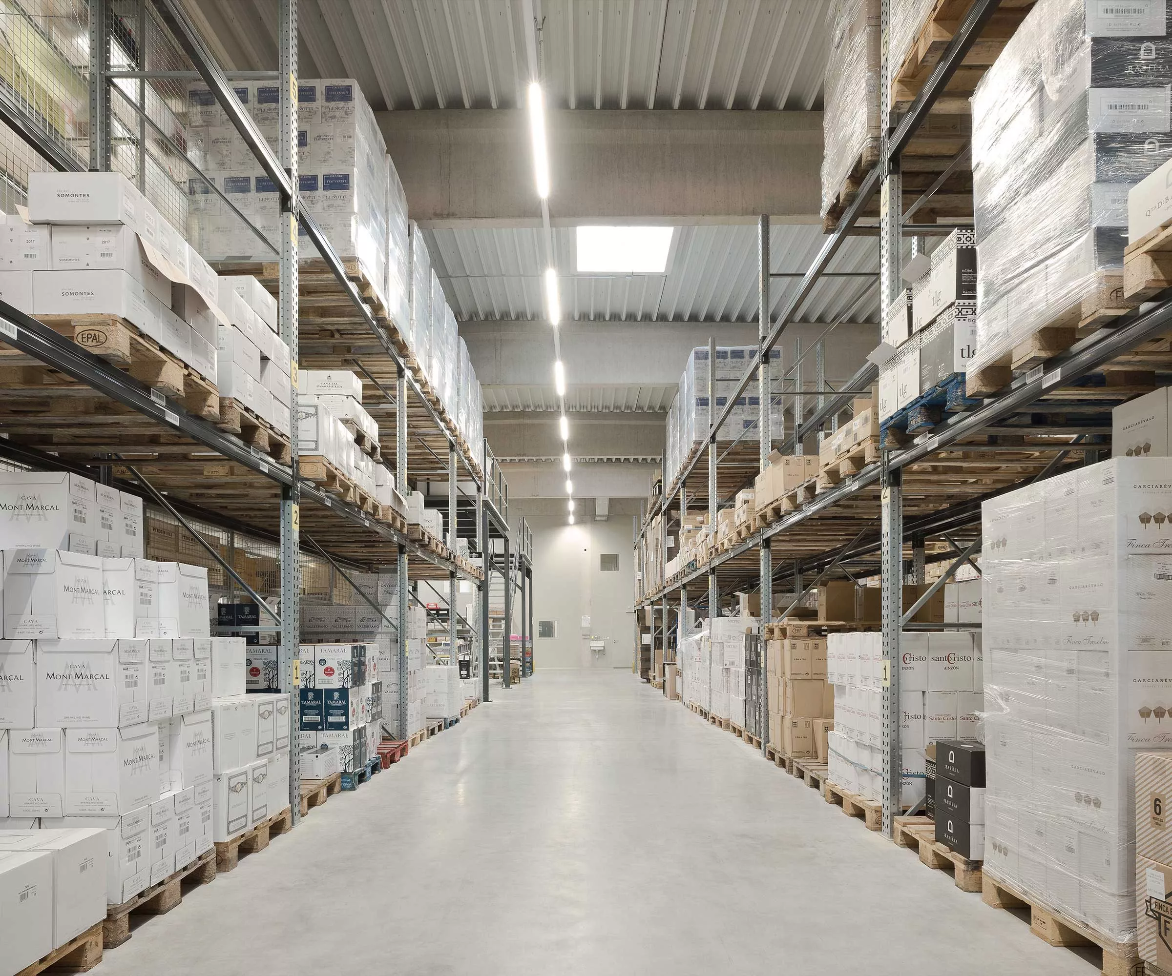Lagerhalle mit hohen Regalen voller Kartons auf Paletten, beleuchtet, leerer Betonboden.
