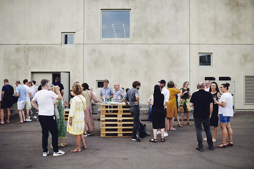 club-traube_Galerie_01 Gruppe von Menschen unterhält sich draußen vor einem Gebäude, um einen Tisch aus Paletten, bei einer Veranstaltung.