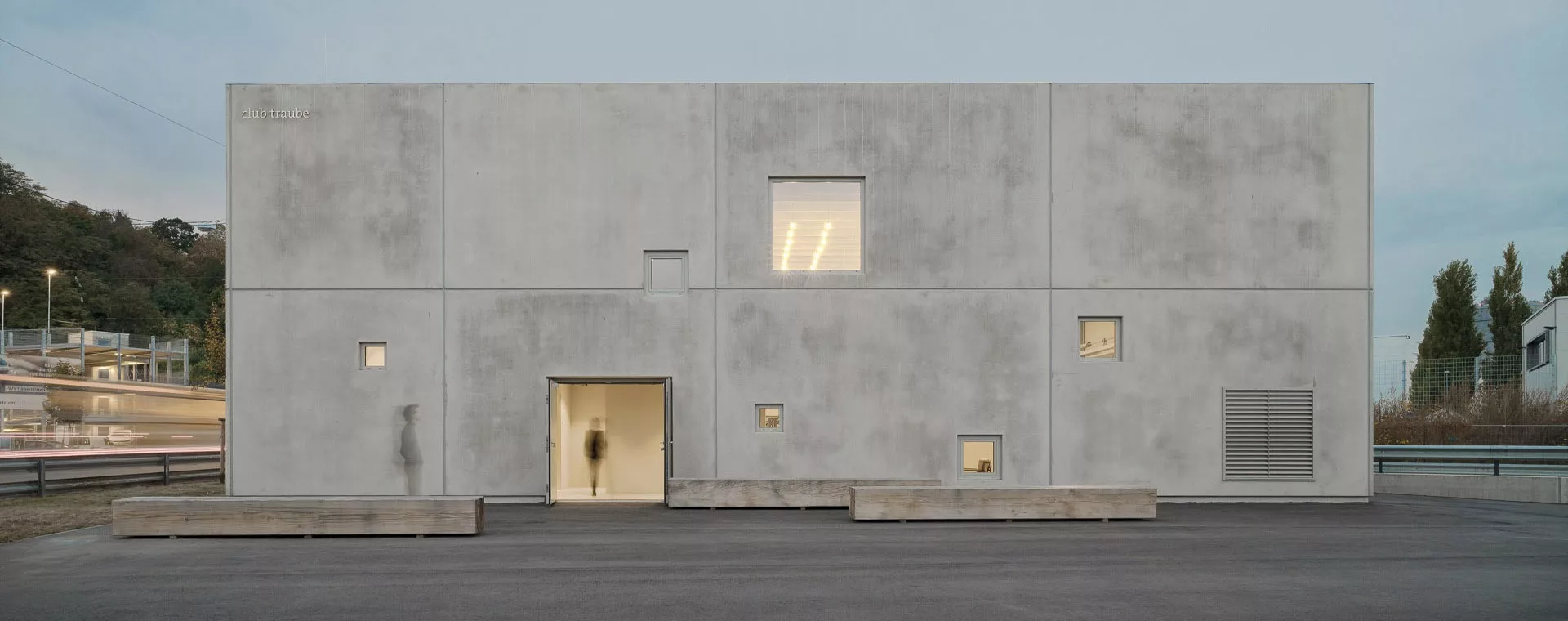 Ein schlichtes, modernes Gebäude mit Betonfassade, kleinen Fenstern und einem Eingang. Menschen sind teilweise unscharf sichtbar.