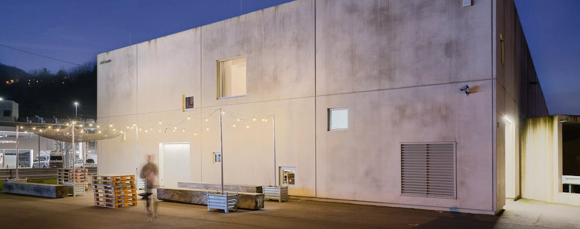 Flaches Gebäude bei Nacht, beleuchtet, eine unscharfe Person läuft, Lichterketten hängen über Paletten im Hof.