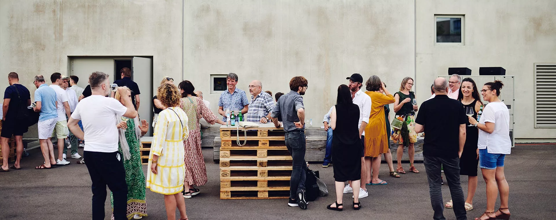 Menschen unterhalten sich und genießen Getränke bei einem informellen Treffen im Freien vor einer schlichten Gebäudewand.
