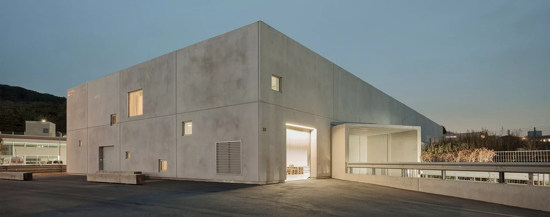 Modernes, beleuchtetes Betongebäude bei Dämmerung, umgeben von Bäumen. Eine offene Tür zeigt Innenbeleuchtung.