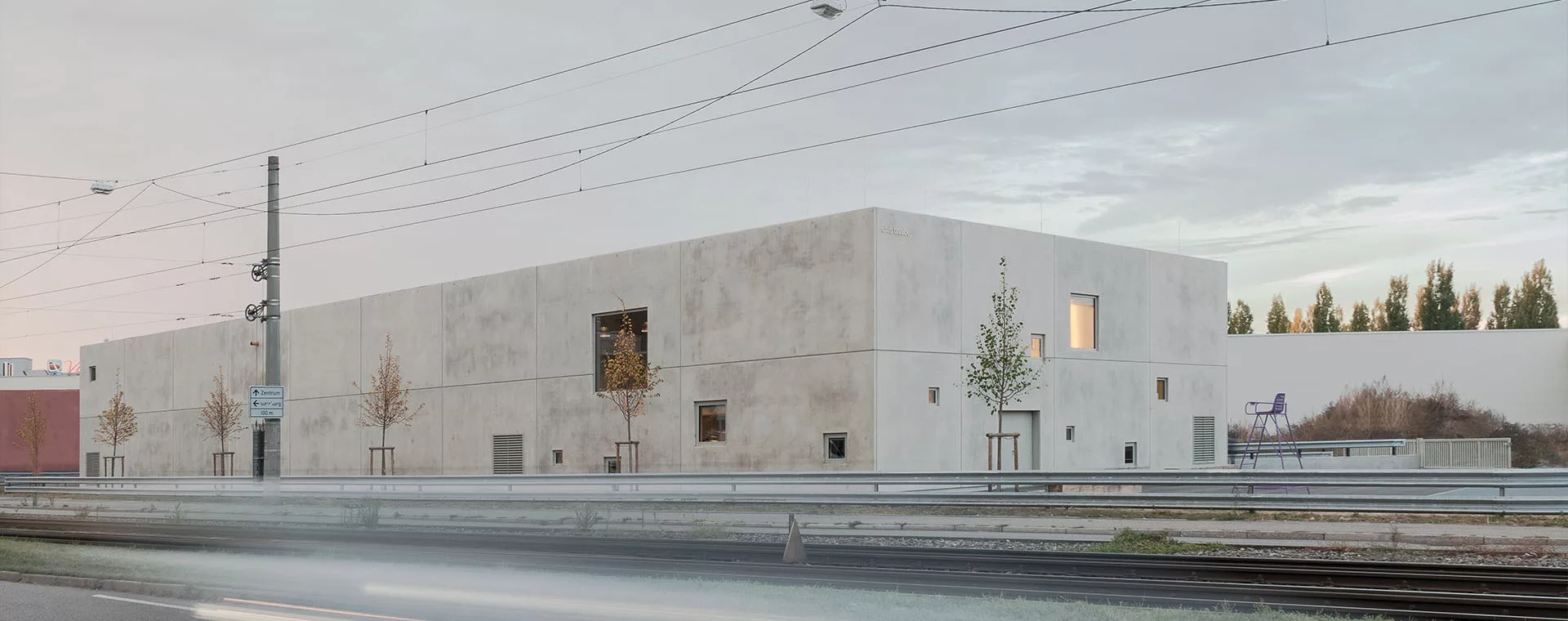 Modernes, rechteckiges Gebäude mit Betonfassade, umgeben von jungen Bäumen, neben einer Straßenbahnlinie bei Sonnenuntergang.