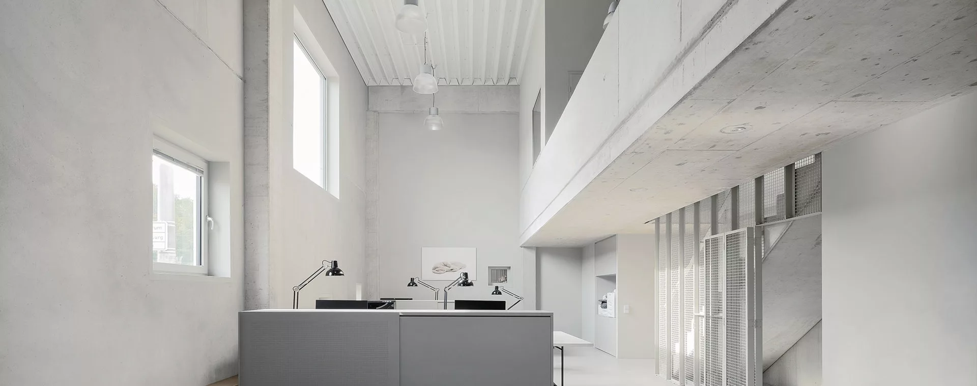 Modernes Büro mit Betonwänden, großen Fenstern und Lampen. Graue Schreibtische mit schwarzen Schreibtischlampen.