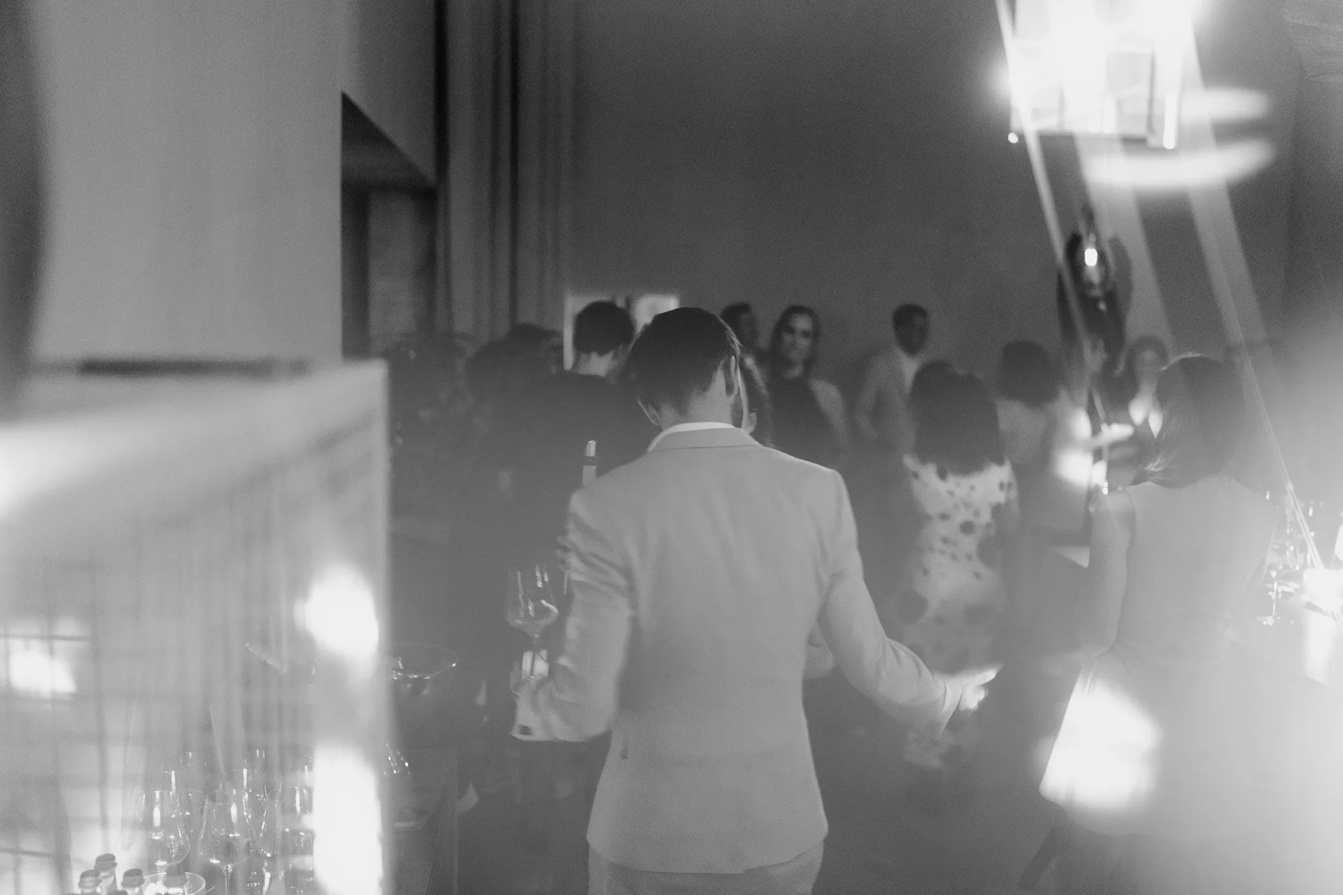 Gruppe von Menschen auf einer Party, teilweise unscharf. Eine Person im Anzug hält ein Weinglas. Stimmung: gesellig, gedämpftes Licht.