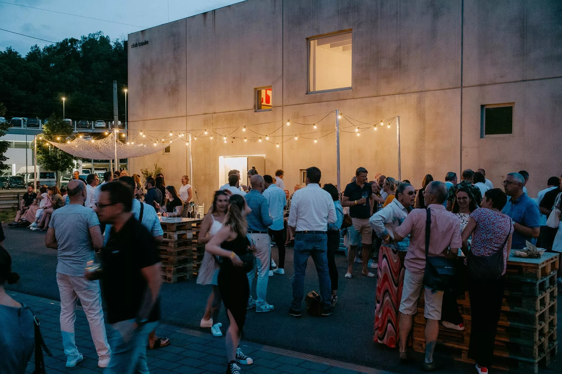 Menschen unterhalten sich bei Abendveranstaltung im Freien vor modernem Gebäude, beleuchtet von Lichterketten.