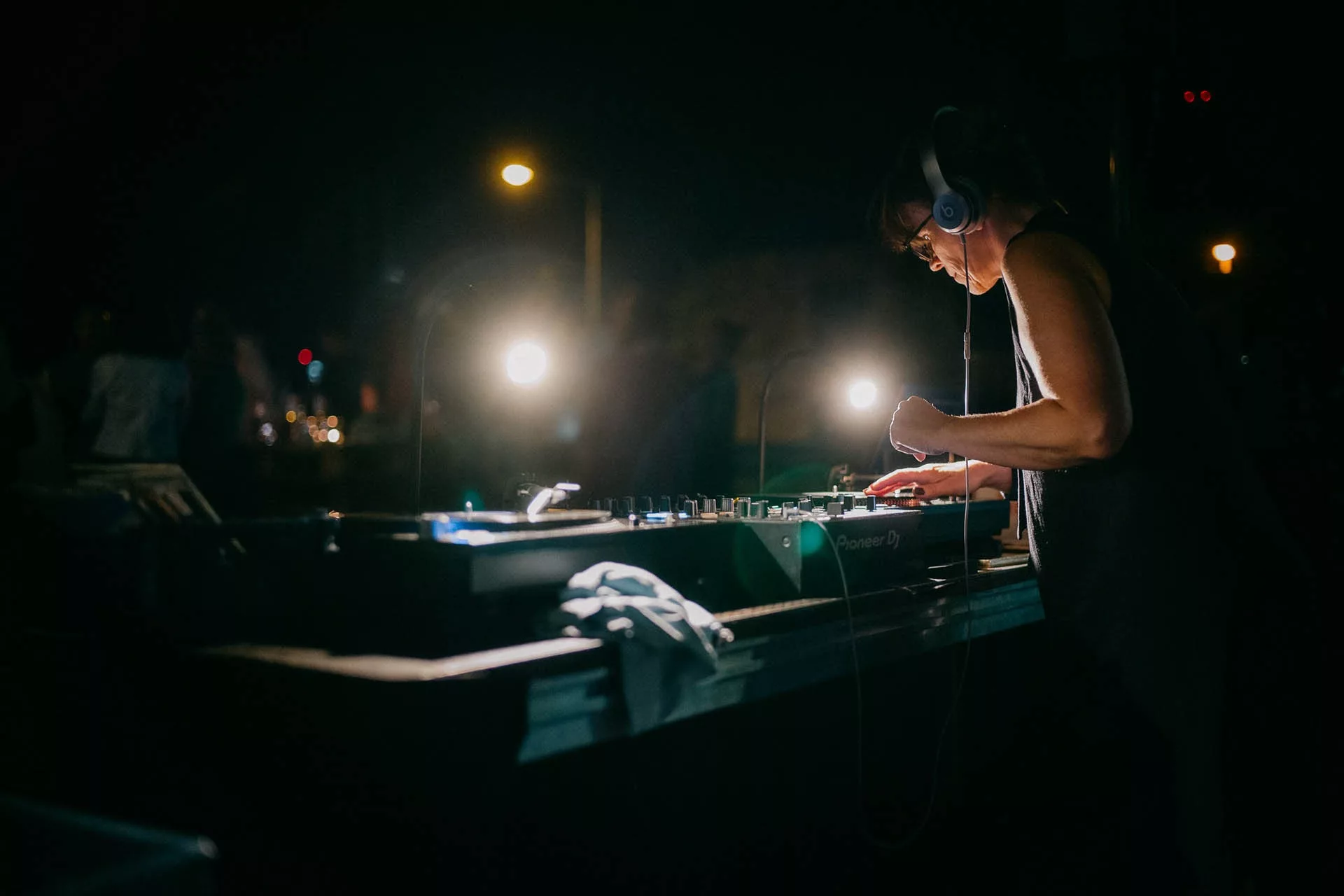 DJ mit Kopfhörern legt nachts im Freien auf, beleuchtet von zwei Lampen. Mischpult und verschwommene Personen im Hintergrund.