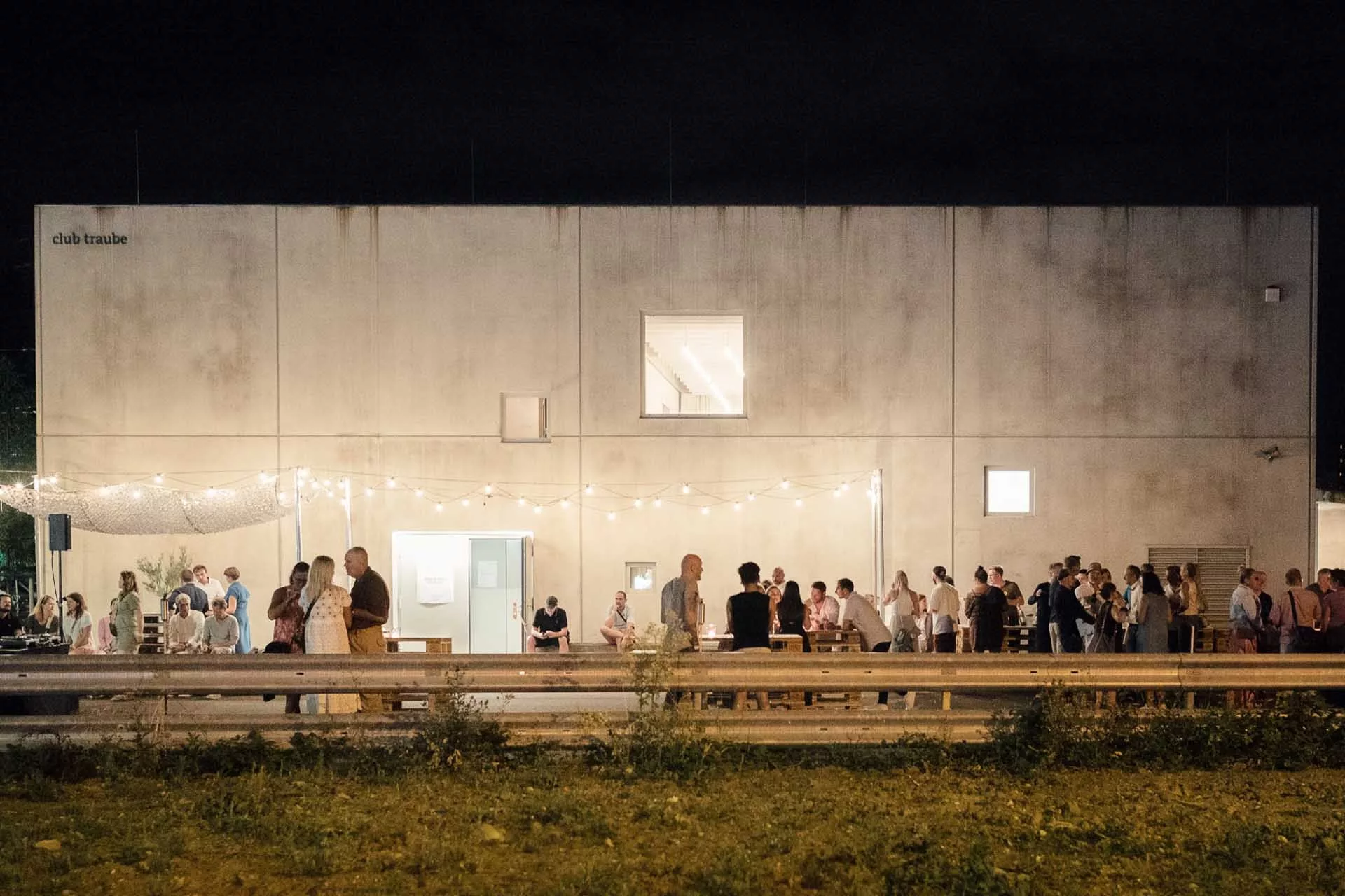 Gruppe von Menschen feiert abends vor einem beleuchteten Gebäude mit Lichterketten, im Freien unter einem dunklen Himmel.