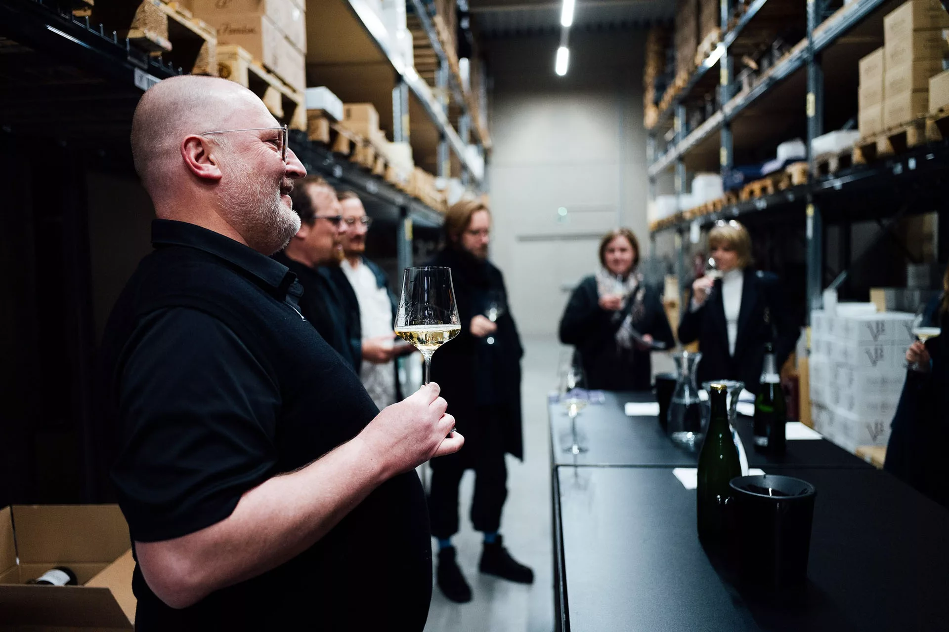 Personen verkosten Wein in einem Lager mit Regalen voller Kisten. Eine Person hält ein Weinglas und lacht.