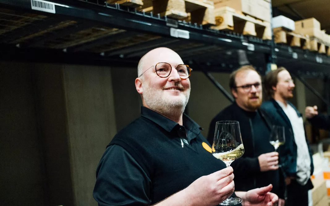Club Traube, Champagner und Co, Deep dive Eine lächelnde Person mit Brille hält ein Glas Weißwein in einem Lagerraum, im Hintergrund weitere Personen mit Gläsern.
