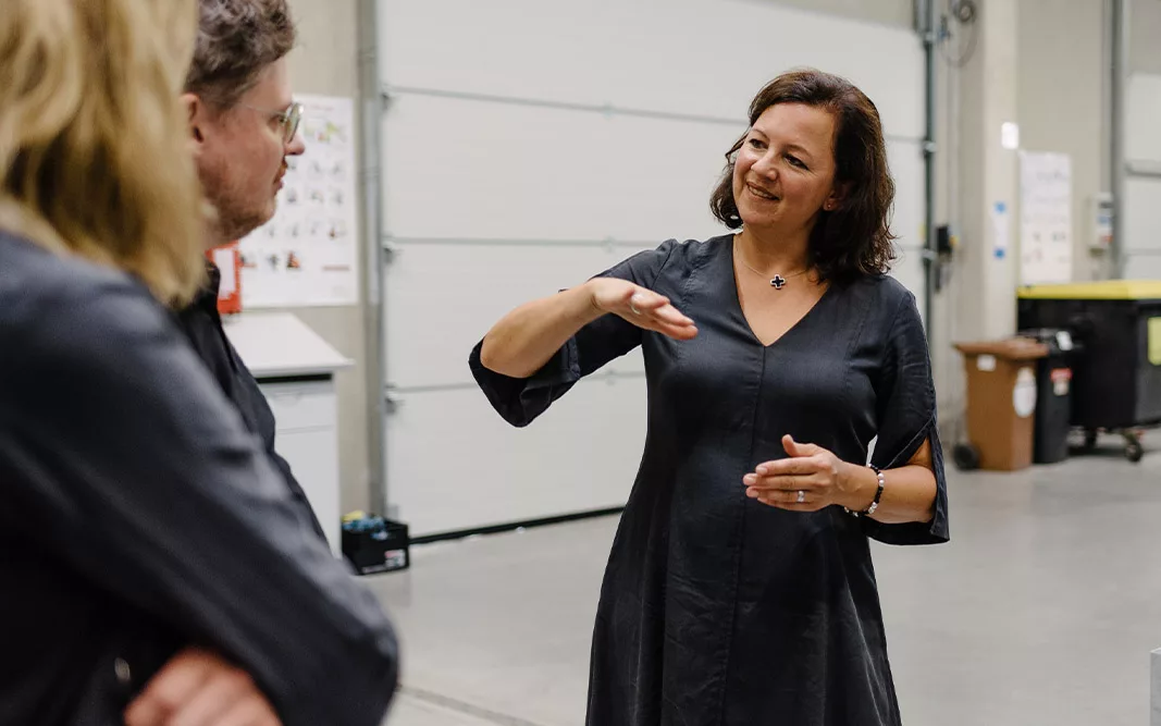 Club Traube, Champagner und Co, Deep dive Eine Frau gestikuliert lächelnd beim Gespräch mit zwei Personen in einer hellen Werkstattumgebung.