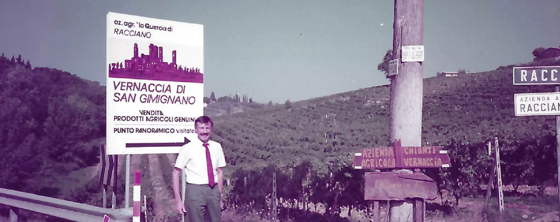 club-traube_ueber-uns_Die-Anfaenge Person vor einem Schild „Vernaccia di San Gimignano” in ländlicher Landschaft, Italien, nahe Racciano.