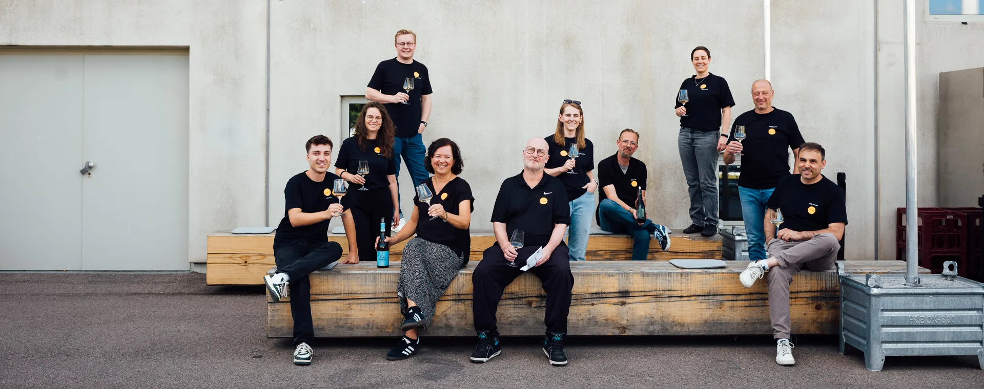 club-traube_ueber-uns_Ein-besonderer-Ort Eine Gruppe von zehn Personen in schwarzen T-Shirts hält Weingläser im Freien. Sie sitzen und stehen auf Holzkisten vor einer Wand.