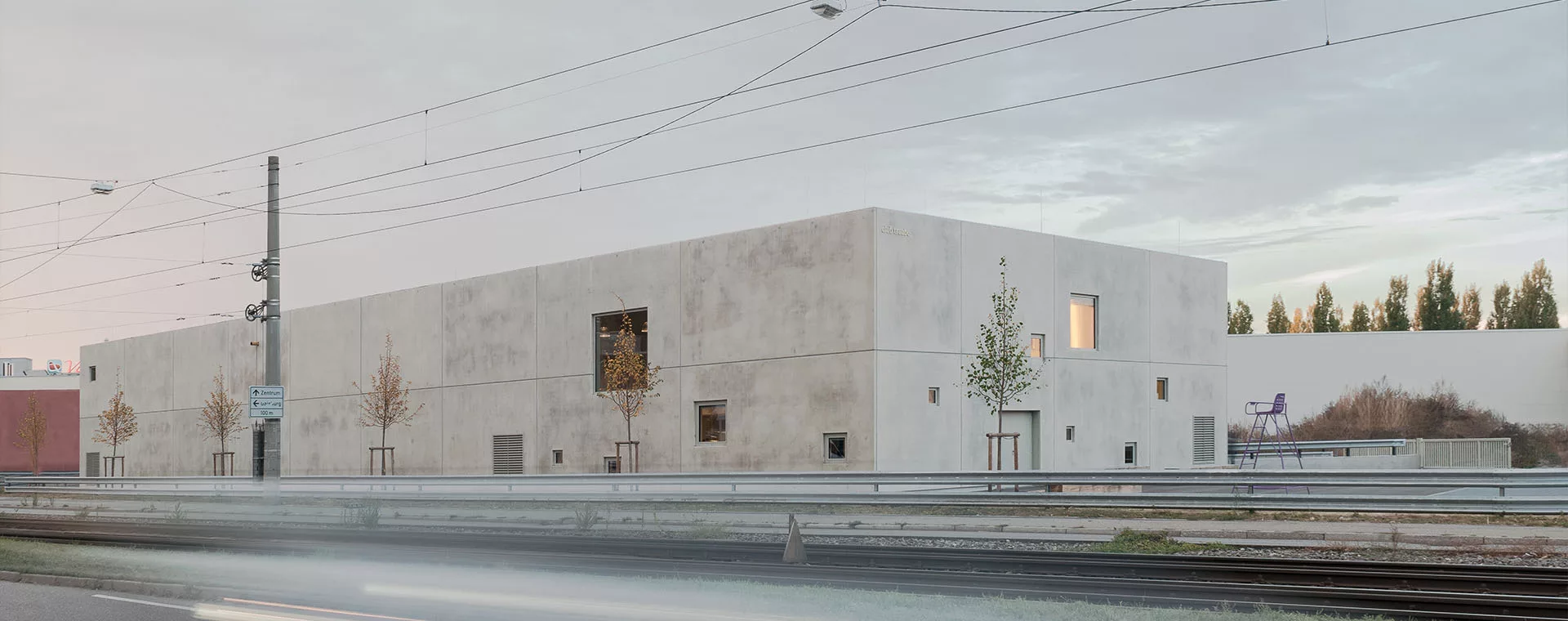 club-traube_ueber-uns_Mehr-als-ein-Standort Modernes, minimalistisches Gebäude aus Beton an einer Straße, umgeben von jungen Bäumen und Gleisen, bei bewölktem Himmel.