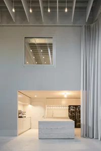 Moderne, minimalistische Küche mit weißem Inseltresen, großem Fenster oben und hohen, grauen Vorhängen in einem loftartigen Raum.